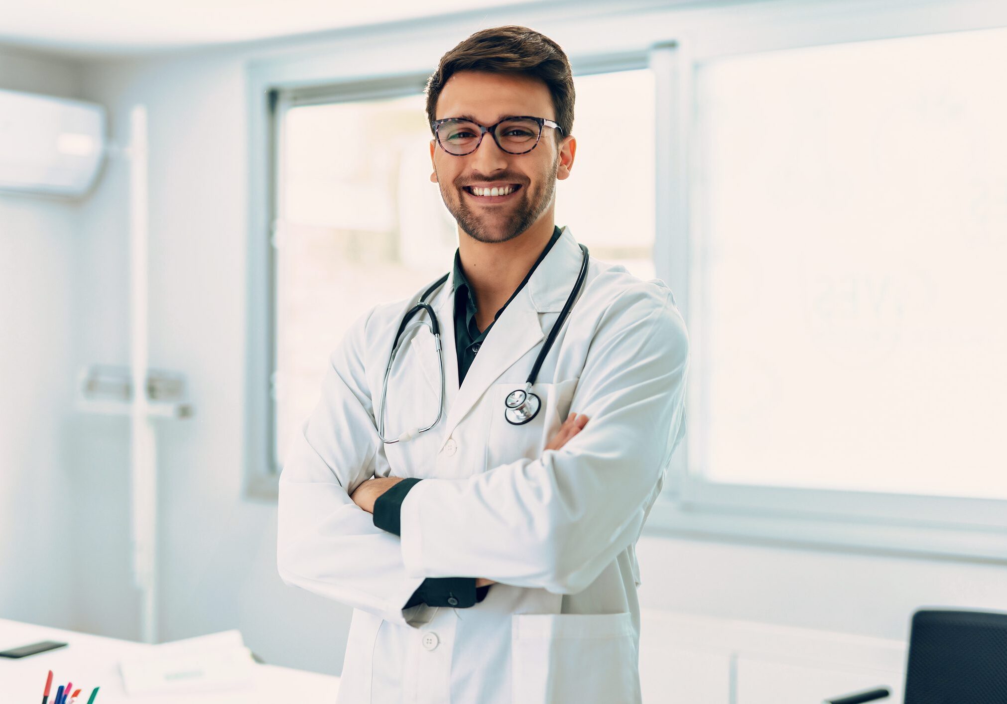 Smiling male doctor with arms crossed in a bright medical office.